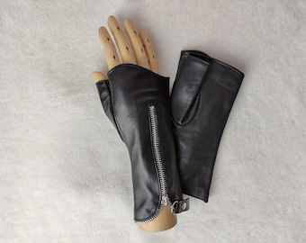 May include: A pair of black leather fingerless gloves featuring a silver-toned metal zipper running vertically along the outer side of the wrist. The gloves have a smooth finish and a small snap button closure at the base of the zipper.