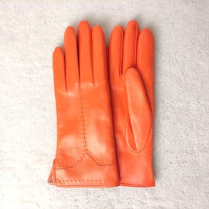 Puede incluir: Un par de guantes de cuero naranja brillante. Los guantes se muestran sobre una superficie blanca texturizada. Tienen un dise&ntilde;o cl&aacute;sico con costuras visibles y una forma ajustada.
