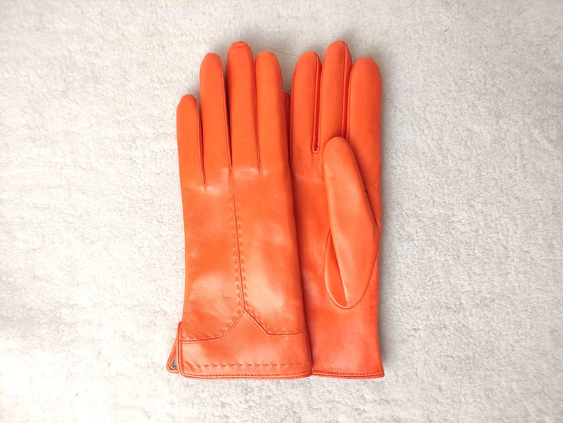 Puede incluir: Un par de guantes de cuero naranja brillante. Los guantes se muestran sobre una superficie blanca texturizada. Tienen un dise&ntilde;o cl&aacute;sico con costuras visibles y una forma ajustada.