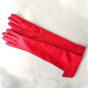 May include: A pair of long, vibrant red leather gloves featuring a smooth finish and a wrist cuff detail secured with two matching red buttons. The gloves extend past the wrist, providing full coverage for the hands and forearms.