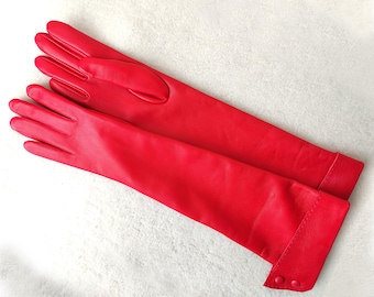 Op de afbeelding: Een paar lange, felrode leren handschoenen met een gladde afwerking en een manchet-detail dat is vastgezet met twee bijpassende rode knopen. De handschoenen reiken tot voorbij de pols en bedekken de handen en onderarmen.
