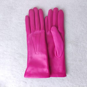 Peut inclure: Une paire de gants en cuir rose vif avec une finition lisse, quatre doigts et un pouce sur chaque main. Le dos des gants pr&eacute;sente trois lignes de couture verticales s'&eacute;tendant de la base des doigts vers le poignet.