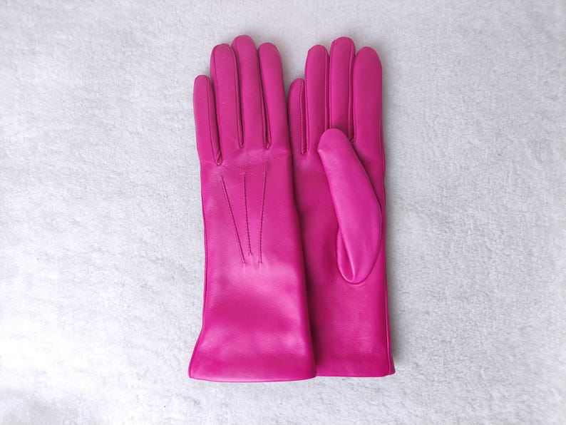 Peut inclure: Une paire de gants en cuir rose vif avec une finition lisse, quatre doigts et un pouce sur chaque main. Le dos des gants pr&eacute;sente trois lignes de couture verticales s'&eacute;tendant de la base des doigts vers le poignet.
