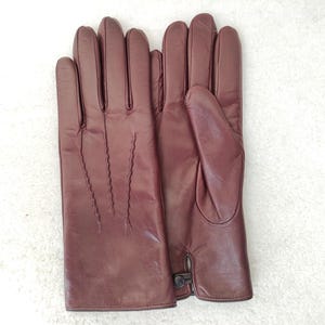May include: A pair of dark burgundy leather gloves featuring three decorative stitched lines on the back of the hand and a small button closure at the wrist. The gloves have a smooth finish and are positioned side by side.