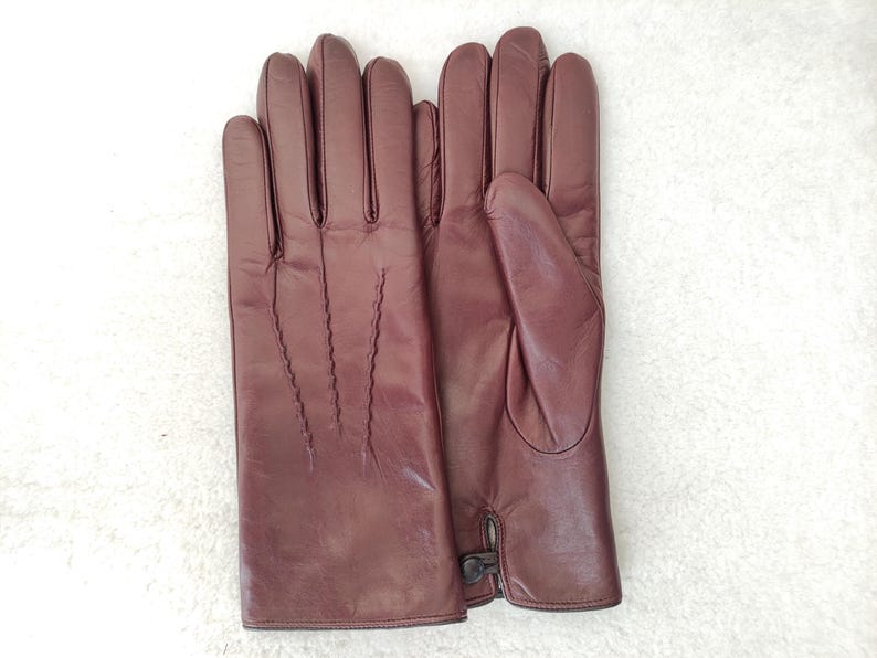 May include: A pair of dark burgundy leather gloves featuring three decorative stitched lines on the back of the hand and a small button closure at the wrist. The gloves have a smooth finish and are positioned side by side.