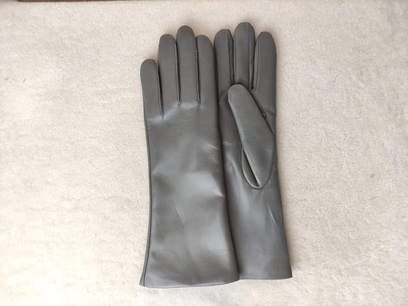 May include: A pair of smooth, matte gray leather gloves with a classic cut. The gloves feature a long wrist design, clean stitching along the fingers, and a simple, unadorned finish. The material has a consistent, solid gray color throughout.