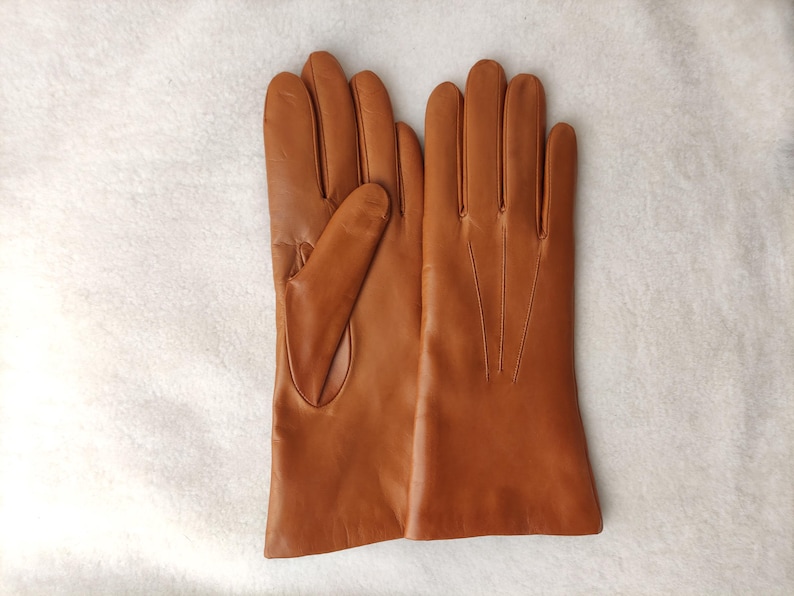 Peut inclure: Une paire de gants en cuir marron clair avec une finition lisse, quatre doigts et un pouce. Le dos du gant droit pr&eacute;sente trois lignes de couture verticales s'&eacute;tendant de la base des doigts vers la zone du poignet.
