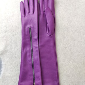 May include: A pair of long, vibrant purple leather gloves featuring a vertical silver-toned metal zip running along the outer forearm of the left glove. The gloves have a smooth finish and extend past the wrist to the mid-forearm area.
