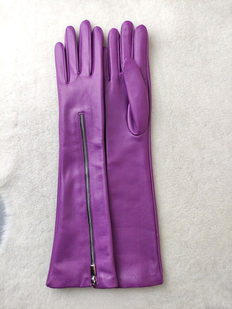 May include: A pair of long, vibrant purple leather gloves featuring a vertical silver-toned metal zip running along the outer forearm of the left glove. The gloves have a smooth finish and extend past the wrist to the mid-forearm area.