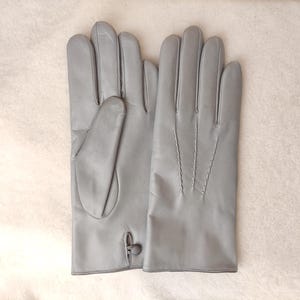 May include: A pair of light gray leather gloves featuring a smooth finish. One glove displays a small button closure at the wrist, while the other glove shows three decorative stitched lines running vertically along the back of the hand.