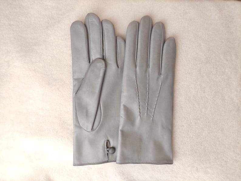 May include: A pair of light gray leather gloves featuring a smooth finish. One glove displays a small button closure at the wrist, while the other glove shows three decorative stitched lines running vertically along the back of the hand.