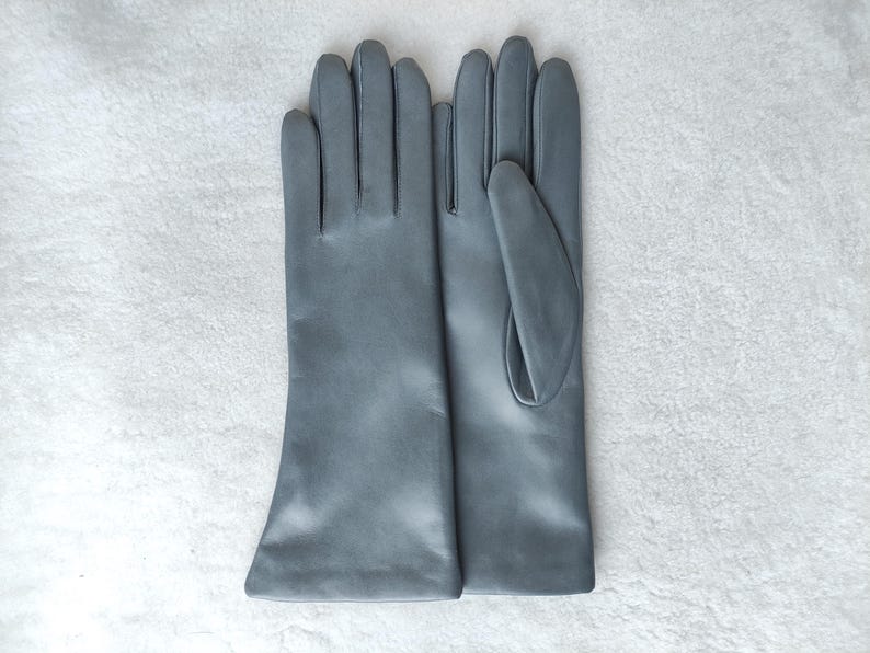 May include: A pair of smooth, matte gray gloves with a classic five-finger design. The gloves feature a clean, seamless construction with a straight wrist opening, showcasing a uniform cool-toned gray color throughout the entire surface.