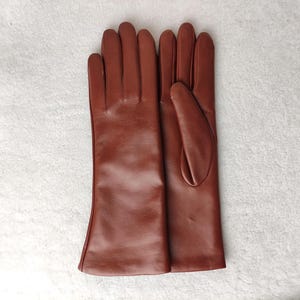 May include: A pair of smooth, medium-brown leather gloves with a classic five-finger design. The gloves feature a clean, minimalist construction with visible stitching along the fingers and a straight, unadorned cuff that extends past the wrist.