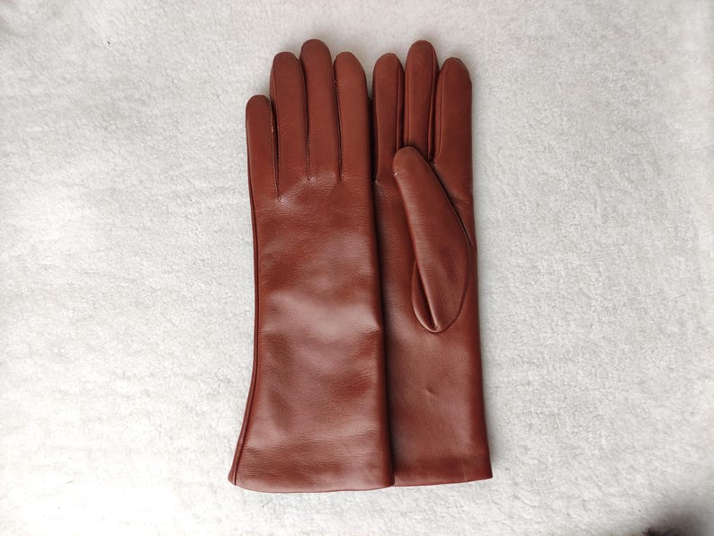 May include: A pair of smooth, medium-brown leather gloves with a classic five-finger design. The gloves feature a clean, minimalist construction with visible stitching along the fingers and a straight, unadorned cuff that extends past the wrist.