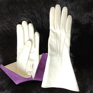 May include: A pair of off-white leather gloves featuring a flared cuff design with a contrasting purple suede interior lining. The gloves include a small button closure at the wrist and decorative stitching details along the back of the hand.