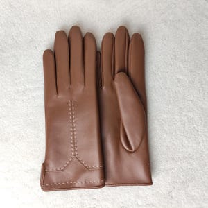 Puede incluir: Un par de guantes de cuero marr&oacute;n. Los guantes son de color marr&oacute;n c&aacute;lido y tienen un dise&ntilde;o cl&aacute;sico. Las costuras son visibles en el dorso de la mano y alrededor de la mu&ntilde;eca. Los guantes son probablemente para el fr&iacute;o.