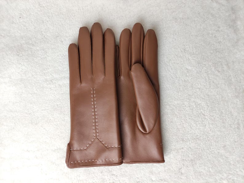 Puede incluir: Un par de guantes de cuero marr&oacute;n. Los guantes son de color marr&oacute;n c&aacute;lido y tienen un dise&ntilde;o cl&aacute;sico. Las costuras son visibles en el dorso de la mano y alrededor de la mu&ntilde;eca. Los guantes son probablemente para el fr&iacute;o.