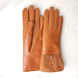May include: A pair of tan leather gloves with a suede cuff. The gloves are a warm brown color and appear to be new. The cuff has a decorative teardrop-shaped cutout.