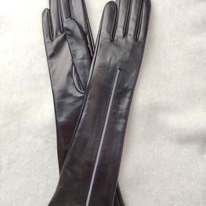 May include: A pair of long black leather gloves featuring a vertical silver-toned metal zip running along the outer forearm of each glove. The gloves have a smooth finish and extend past the wrist, with a visible zip pull at the cuff.
