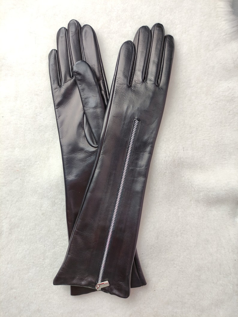May include: A pair of long black leather gloves featuring a vertical silver-toned metal zip running along the outer forearm of each glove. The gloves have a smooth finish and extend past the wrist, with a visible zip pull at the cuff.
