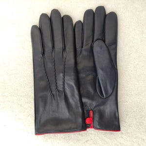 May include: A pair of dark navy blue leather gloves featuring three decorative stitched lines on the back of the hand and a small red button detail at the wrist opening, with a thin red trim along the bottom edge of the cuffs.