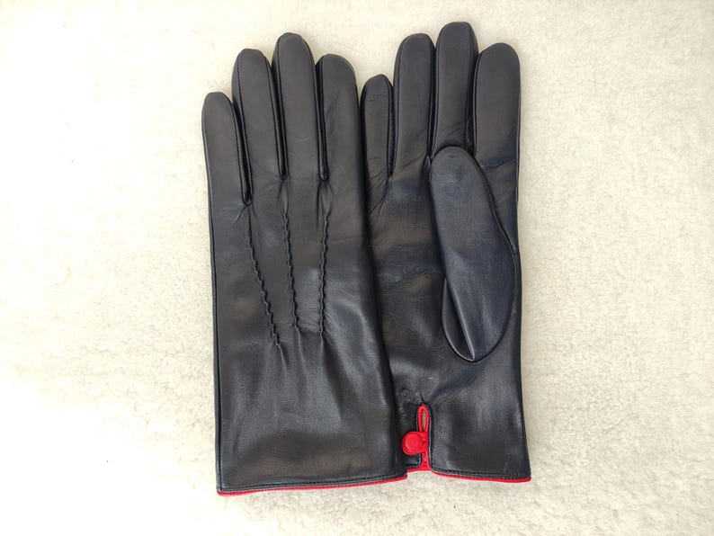 May include: A pair of dark navy blue leather gloves featuring three decorative stitched lines on the back of the hand and a small red button detail at the wrist opening, with a thin red trim along the bottom edge of the cuffs.