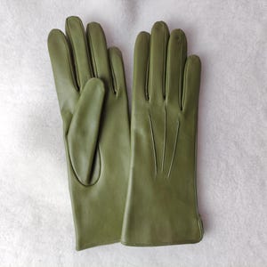 Peut inclure: Une paire de gants en cuir vert olive avec une finition lisse, quatre doigts et un pouce. Le dos des gants pr&eacute;sente trois lignes verticales cousues s'&eacute;tendant du poignet vers les articulations pour un d&eacute;tail suppl&eacute;mentaire.