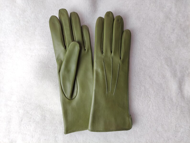Peut inclure: Une paire de gants en cuir vert olive avec une finition lisse, quatre doigts et un pouce. Le dos des gants pr&eacute;sente trois lignes verticales cousues s'&eacute;tendant du poignet vers les articulations pour un d&eacute;tail suppl&eacute;mentaire.