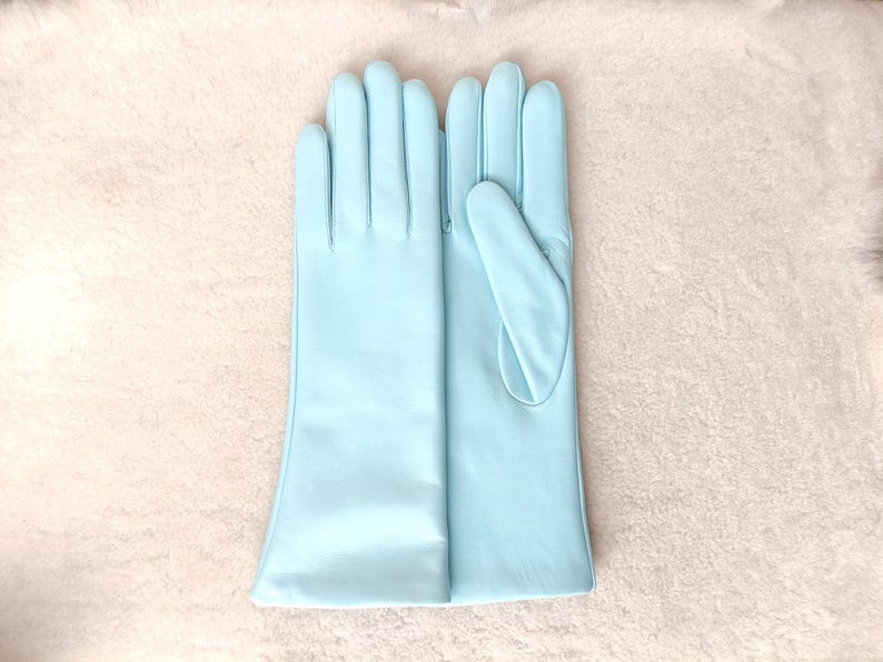 May include: A pair of light blue opera-length gloves made of a smooth, matte material. The gloves feature a classic design with four fingers and a thumb, extending past the wrist to the mid-forearm, showcasing a clean and uniform finish.