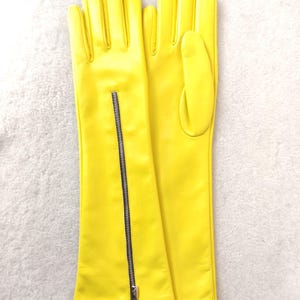 May include: A pair of bright yellow leather gloves featuring a long cuff design. The left glove includes a vertical silver-toned metal zip running from the wrist towards the forearm, while the right glove displays a smooth, plain surface.