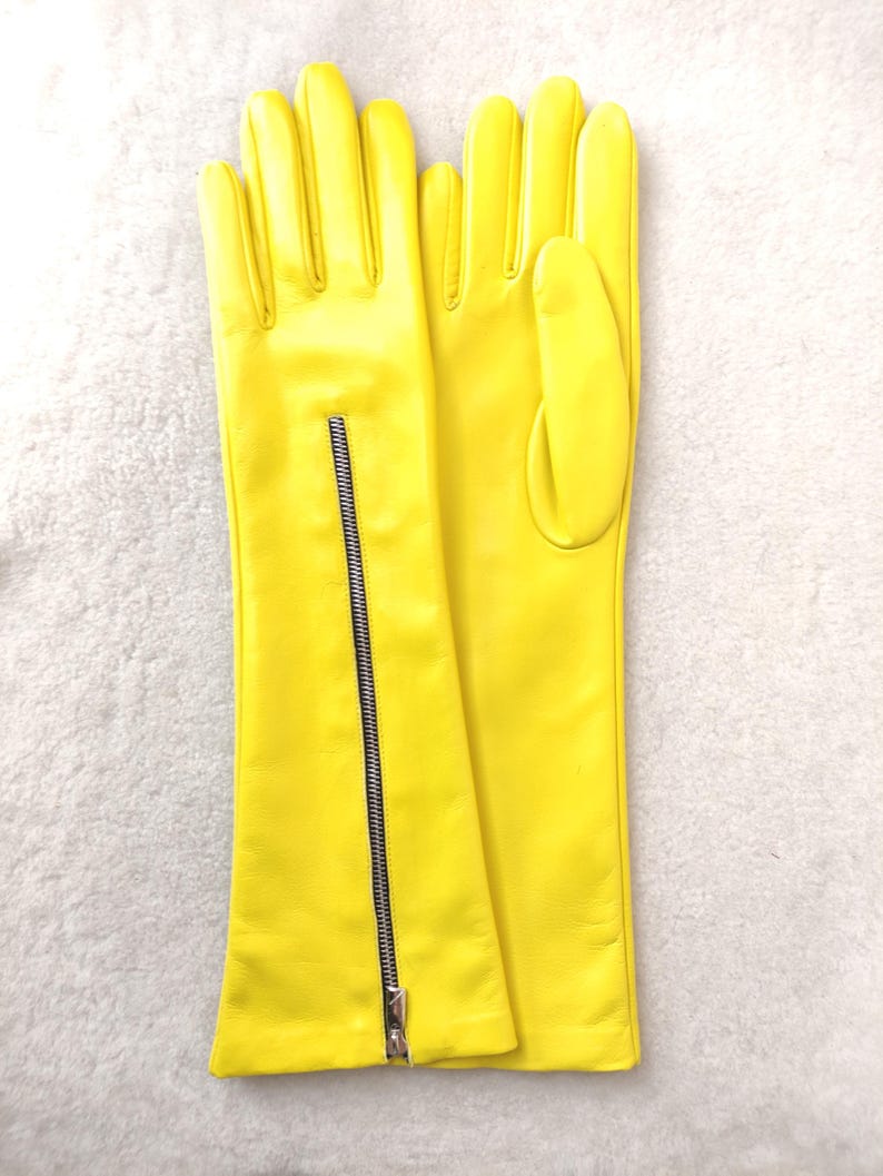 May include: A pair of bright yellow leather gloves featuring a long cuff design. The left glove includes a vertical silver-toned metal zip running from the wrist towards the forearm, while the right glove displays a smooth, plain surface.