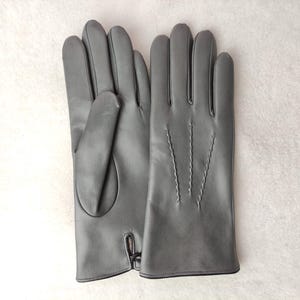May include: A pair of smooth grey gloves featuring a classic design with three decorative stitched lines on the back of the hand and a small slit detail at the wrist. The gloves have a clean, uniform finish and a structured shape.