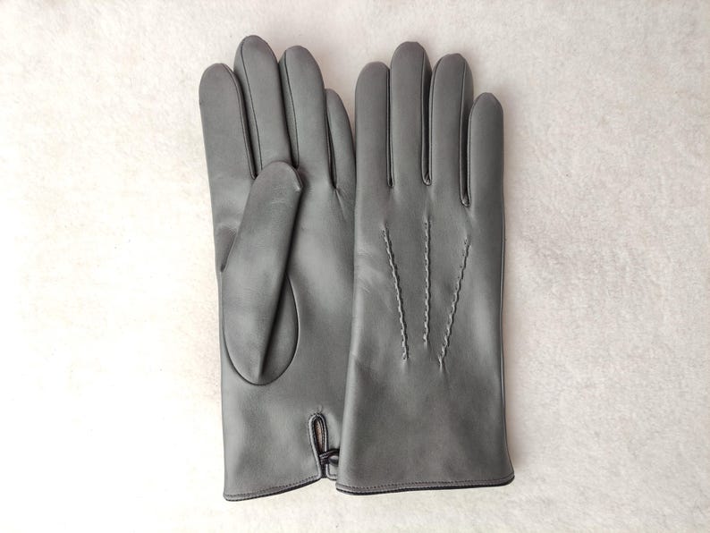 May include: A pair of smooth grey gloves featuring a classic design with three decorative stitched lines on the back of the hand and a small slit detail at the wrist. The gloves have a clean, uniform finish and a structured shape.