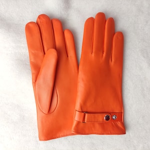 May include: A pair of bright orange leather gloves featuring a smooth finish and a wrist strap detail with a silver-toned metal snap button closure. The gloves are designed with five fingers and a clean, minimalist construction.