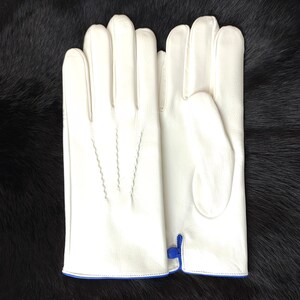 May include: A pair of off-white leather gloves featuring three decorative stitched lines on the back of the hand and a small blue zipper detail at the wrist opening. The gloves are smooth in texture and have a clean, structured design.