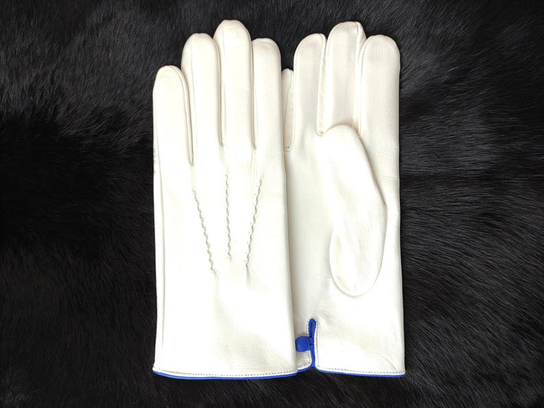 May include: A pair of off-white leather gloves featuring three decorative stitched lines on the back of the hand and a small blue zipper detail at the wrist opening. The gloves are smooth in texture and have a clean, structured design.