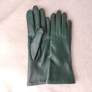 May include: A pair of dark forest green leather gloves with smooth, matte-finish surfaces. The gloves feature a classic five-finger design with stitched detailing along the fingers and a simple, straight-cut wrist opening for a clean look.