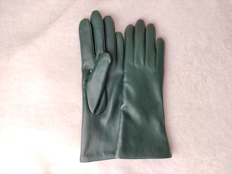 May include: A pair of dark forest green leather gloves with smooth, matte-finish surfaces. The gloves feature a classic five-finger design with stitched detailing along the fingers and a simple, straight-cut wrist opening for a clean look.