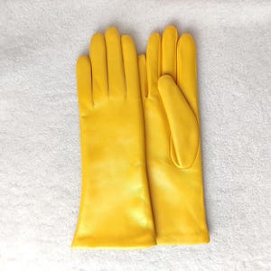 May include: A pair of bright yellow leather gloves with a smooth finish. The gloves feature a classic design with four fingers and a thumb, extending past the wrist. The material has a consistent, vibrant yellow color throughout the surface.