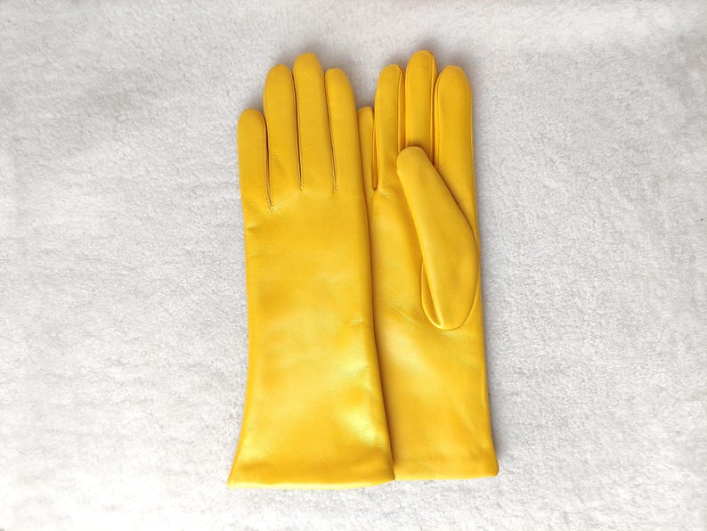 May include: A pair of bright yellow leather gloves with a smooth finish. The gloves feature a classic design with four fingers and a thumb, extending past the wrist. The material has a consistent, vibrant yellow color throughout the surface.