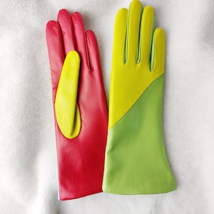 Puede incluir: Un par de guantes de cuero con un diseño de bloques de color. Un guante es principalmente rojo con un acento de pulgar amarillo brillante, mientras que el otro guante está dividido diagonalmente entre una sección superior amarilla y una inferior verde claro.