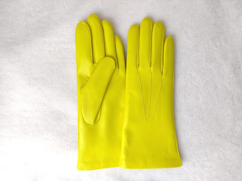 Peut inclure: Une paire de gants jaune n&eacute;on vif avec une finition lisse, quatre doigts et un pouce. Le dos du gant droit pr&eacute;sente trois lignes verticales cousues s'&eacute;tendant de la base des doigts vers la zone du poignet.