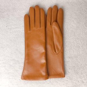 May include: A pair of smooth, tan-colored leather gloves with a classic design. The gloves feature a clean, minimalist aesthetic with visible stitching along the fingers and a simple, straight-cut wrist opening, showcasing a uniform finish.