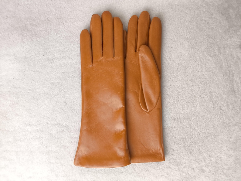 May include: A pair of smooth, tan-colored leather gloves with a classic design. The gloves feature a clean, minimalist aesthetic with visible stitching along the fingers and a simple, straight-cut wrist opening, showcasing a uniform finish.