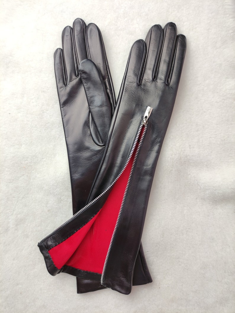 May include: A pair of black leather gloves featuring a silver-toned metal zip running along the side of the wrist. The interior lining of the gloves is a vibrant red fabric, which is visible where the zip is partially opened.