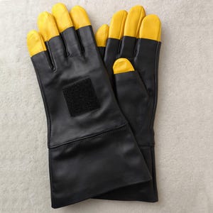 Può includere: Un paio di guanti in pelle nera con le punte delle dita gialle. I guanti hanno un polsino lungo e una toppa quadrata nera sul dorso della mano. Questi guanti sono probabilmente per protezione o lavoro.