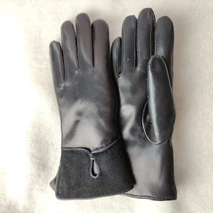 May include: A pair of black leather gloves. The gloves have a smooth, shiny finish on the hand portion and a contrasting dark suede cuff. The cuff features a small, decorative teardrop-shaped cutout. The gloves are laid out on a light-colored surface.