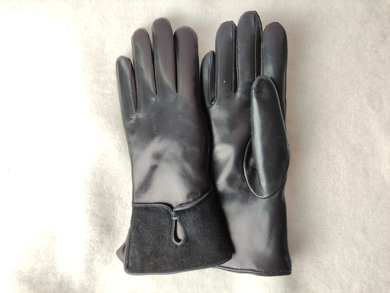 May include: A pair of black leather gloves. The gloves have a smooth, shiny finish on the hand portion and a contrasting dark suede cuff. The cuff features a small, decorative teardrop-shaped cutout. The gloves are laid out on a light-colored surface.