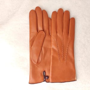 May include: A pair of smooth, cognac-colored leather gloves featuring four vertical decorative stitched lines on the back of the hand and a small, dark brown button closure detail at the wrist opening. The gloves have a clean, polished finish.