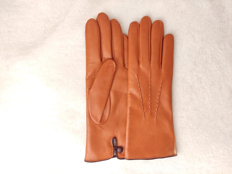 May include: A pair of smooth, cognac-colored leather gloves featuring four vertical decorative stitched lines on the back of the hand and a small, dark brown button closure detail at the wrist opening. The gloves have a clean, polished finish.